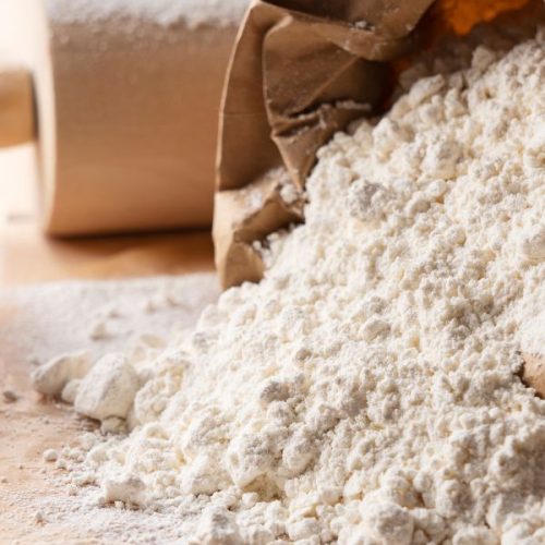 A paper bag spills flour onto a wooden surface, hinting at the creation of ancient grain rolls, with a rolling pin poised in the background.
