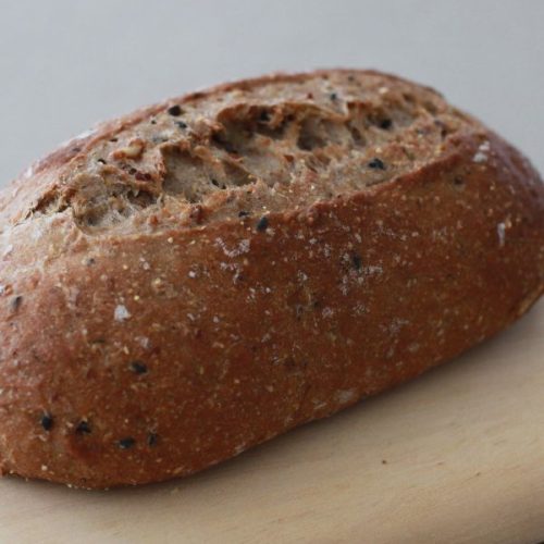 A loaf of brown bread with a rugged crust, reminiscent of a classic farro bread recipe, rests on a light wooden surface.