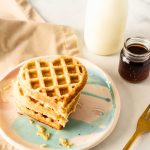 A stack of heart-shaped protein waffles with stevia sits on a colorful plate, alongside a bottle of milk, a jar of syrup, a gold fork, and a beige napkin on a white surface.