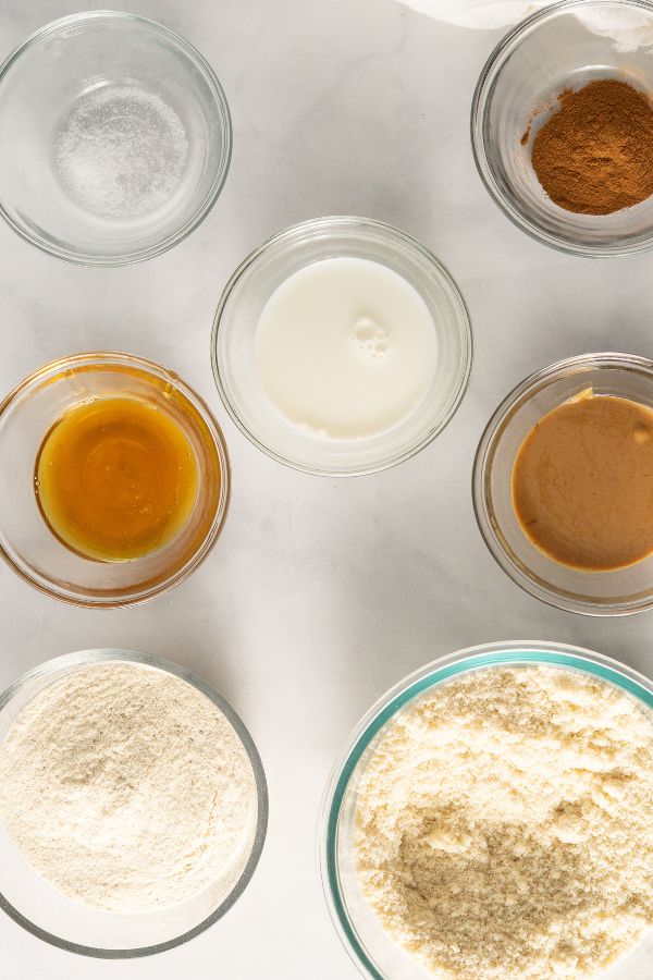 Seven glass bowls containing various baking ingredients for Churro Protein Balls, including flour, almond flour, milk, honey, peanut butter, cinnamon, and salt, are neatly arranged on a white surface.