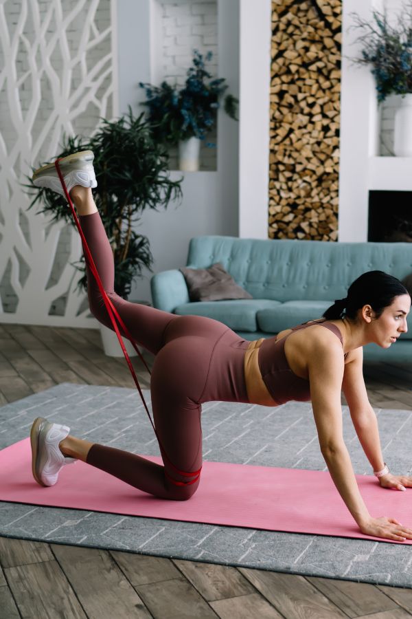 Woman exercising on a pink yoga mat at home, performing Glute Kick Backs with a resistance band in athletic wear.