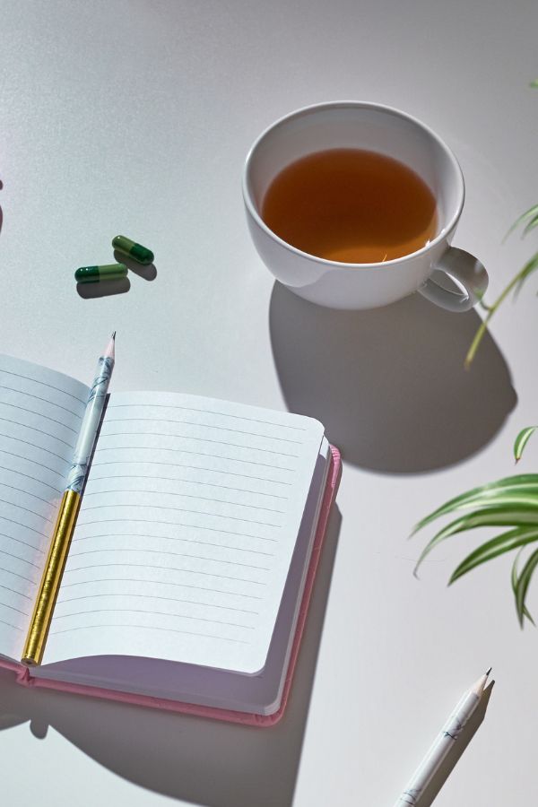 An open lined notebook with a pencil, a cup of tea, two green capsules—perfect for supporting enough quality sleep—and a plant on a white surface.