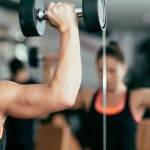 Person performing shoulder presses with dumbbells in front of a mirror at a gym, wearing a black tank top with orange trim.