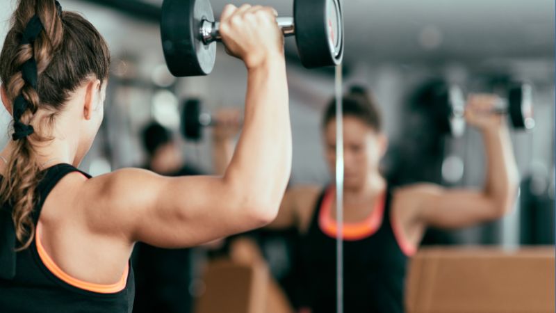 Person performing shoulder presses with dumbbells in front of a mirror at a gym, wearing a black tank top with orange trim.