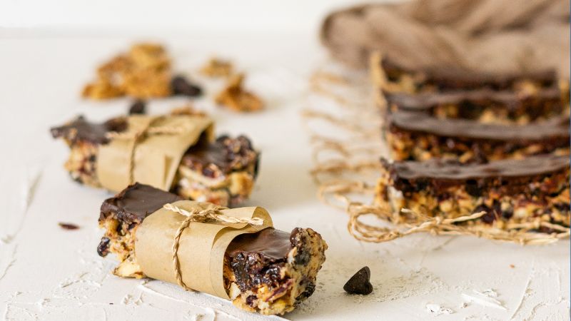 Four chocolate-topped Kind Bars, two wrapped in brown paper, are placed on a white surface with scattered granola pieces and chocolate chips in the background.