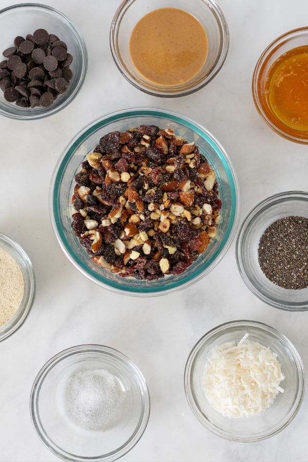 Several glass bowls containing ingredients like chocolate chips, nut butter, honey, chopped nuts and dried fruit, chia seeds, shredded coconut, protein powder, and salt are arranged on a white surface—perfect for making homemade snacks inspired by Kind Bars.