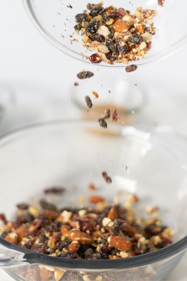 A bowl of mixed nuts and dried fruit, like those found in Kind Bars, is being poured into another clear glass bowl, with some ingredients captured falling mid-air.