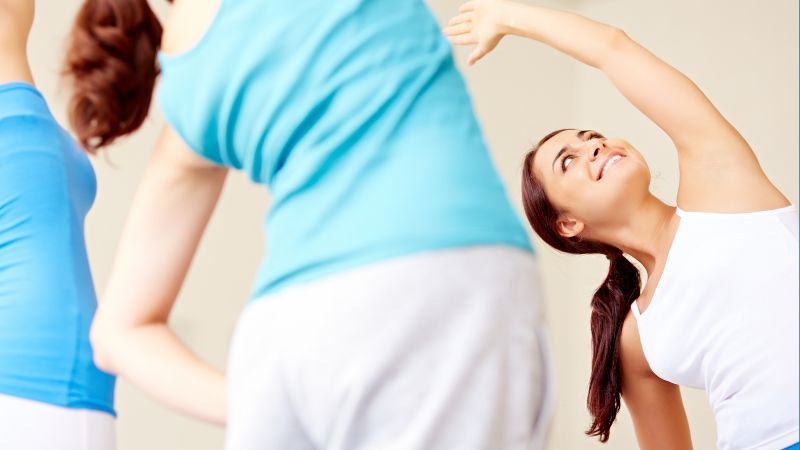 Three people in athletic wear participate in a group exercise class, with one woman smiling as she performs Side Bends and stretches to the side.
