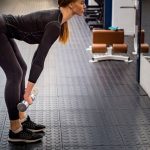 Woman in athletic wear demonstrates how to deadlift with dumbbells, performing the move in front of a mirror on a black rubber gym floor.