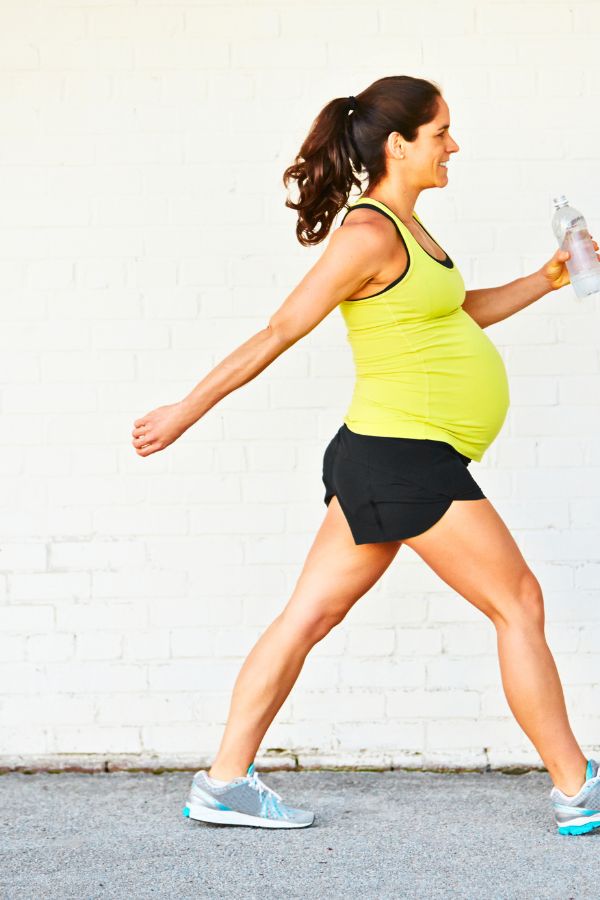 Pregnant woman in athletic wear briskly walking outdoors, holding a water bottle and smiling, with a white brick wall in the background—demonstrating one of the safe exercises during pregnancy.