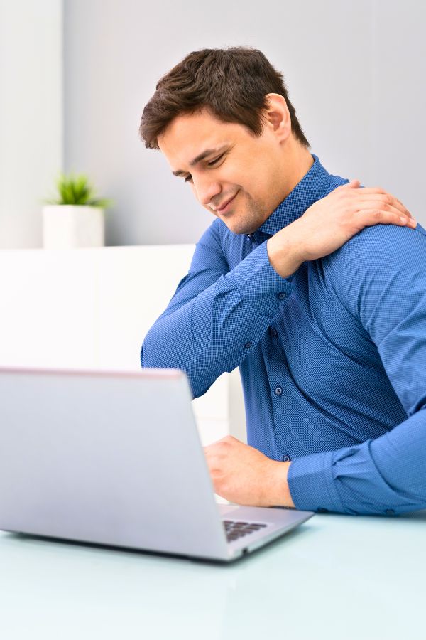 A man in a blue shirt sits at a desk with a laptop, holding his left shoulder and appearing to feel discomfort, possibly from trigger points.