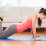 A woman in athletic wear performs modified push-ups, a popular strength training exercise, on a blue mat in a living room with a couch in the background.