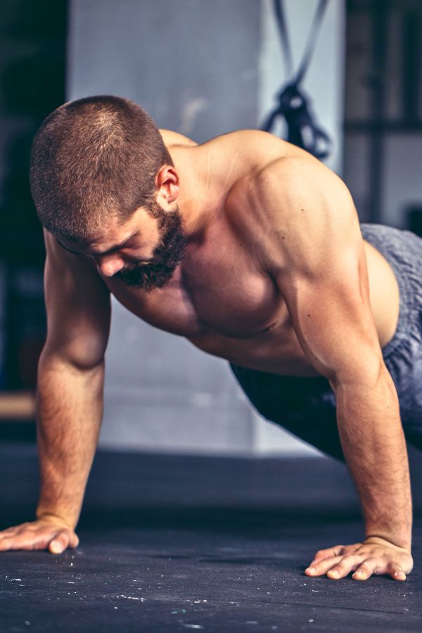 A shirtless man with a beard performs a push-up on a gym floor, focusing downward, his muscles visibly engaged in one of many strength training exercises.