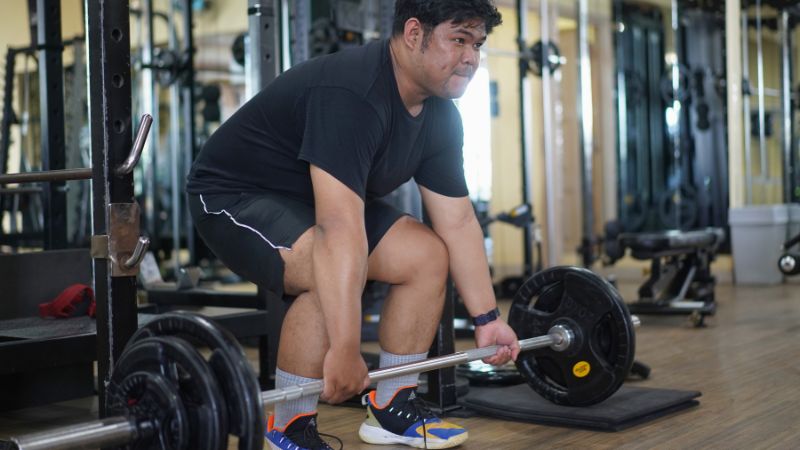 A man in athletic wear prepares to lift a loaded barbell in a gym, surrounded by various exercise equipment, demonstrating proper form for how to deadlift.