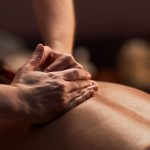 A person receives a back massage for stress and anxiety from another individual, with hands applying pressure to the upper back and shoulders in a dimly lit setting.