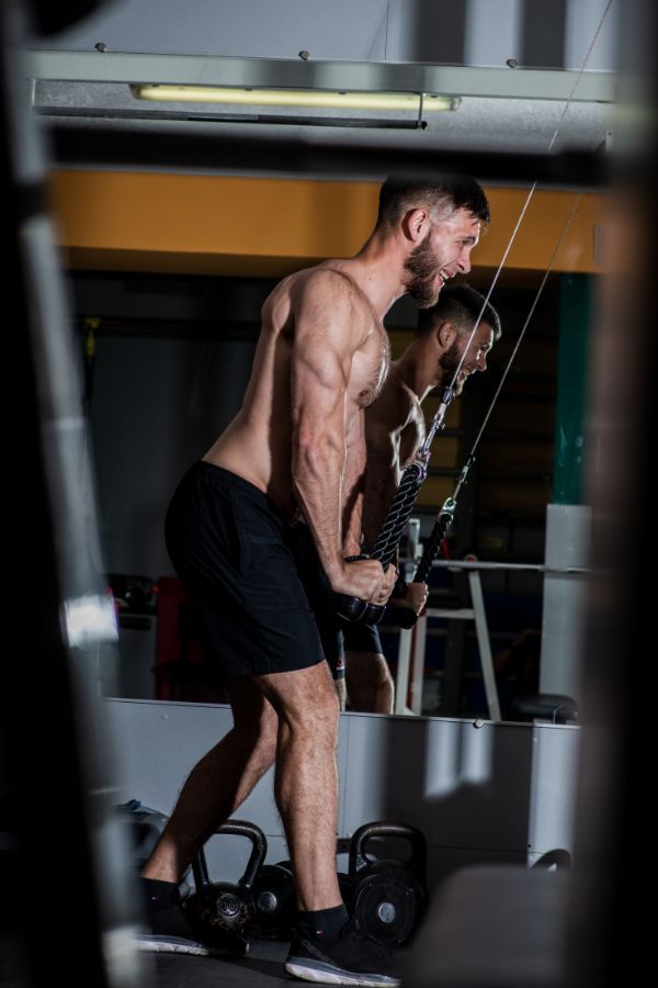 A shirtless man in black shorts performs tricep extensions using a cable machine in a gym, with kettlebells on the floor and his reflection visible in the mirror.