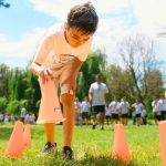 Children participate in an outdoor relay race, moving orange cones on a grassy field with trees and other kids in the background—perfect Family Exercises for Kids that promote teamwork and active fun.