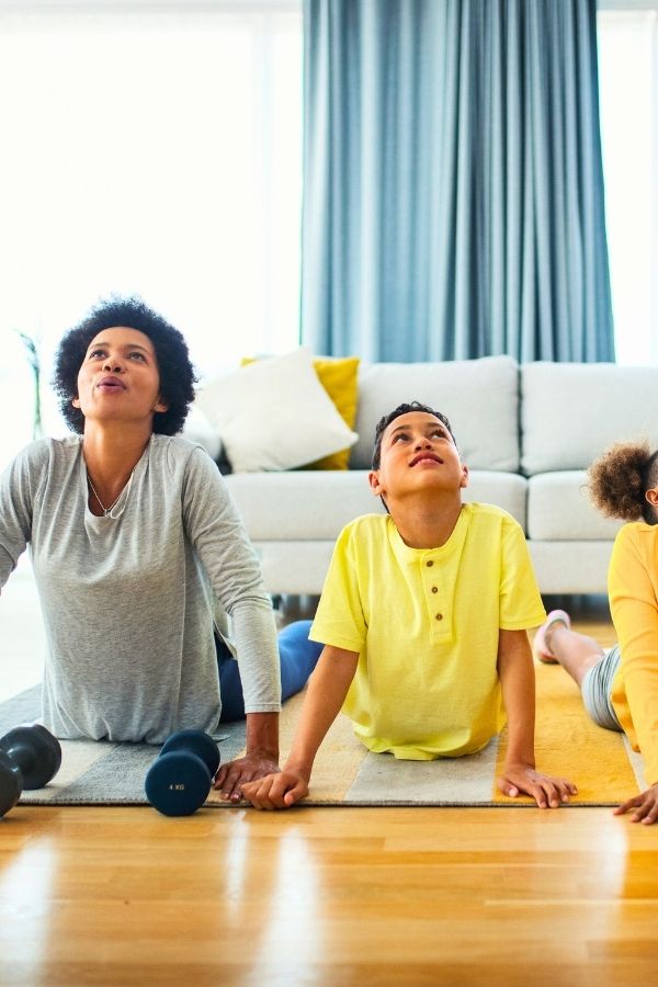 A family enjoys a yoga session in their living room, with an adult and two children holding a cobra pose on a rug. Dumbbells rest nearby, making this scene a perfect example of Family Exercises for Kids.