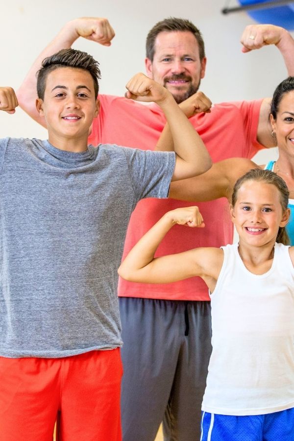 Two kids and two adults stand together indoors, smiling and flexing their arm muscles, showcasing strength and confidence—perfectly capturing the spirit of family exercises for kids.