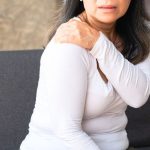 A woman sitting on a couch holds her left shoulder with her right hand, appearing to experience shoulder pain, possibly in need of gentle exercises for osteoporosis.