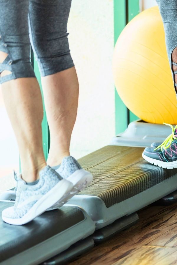 Two people wearing athletic shoes step onto aerobic steppers in a gym, demonstrating exercises for osteoporosis, with a yellow exercise ball visible in the background.