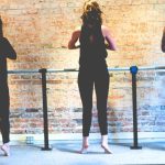 Three people in workout clothes stand on tiptoes facing a barre against a brick wall, appearing to perform calf raises as part of a fitness or barre class.