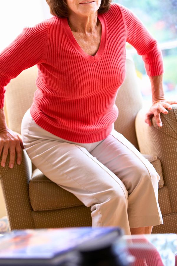 An older woman wearing a red sweater and beige pants is performing chair squats, standing up from a beige armchair with her hands on the armrests for support.