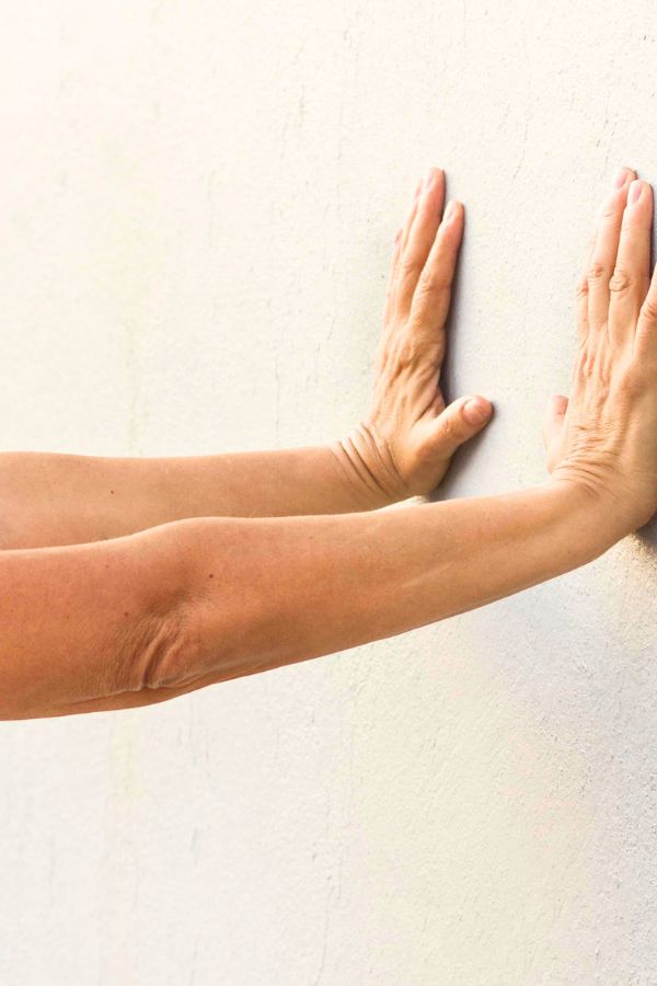 Person performing Wall Push Ups by pressing both hands against a white wall, arms extended and palms flat on the surface.