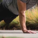 A person wearing a gray shirt and black shorts is doing a Push Ups Workout outdoors on a concrete surface.