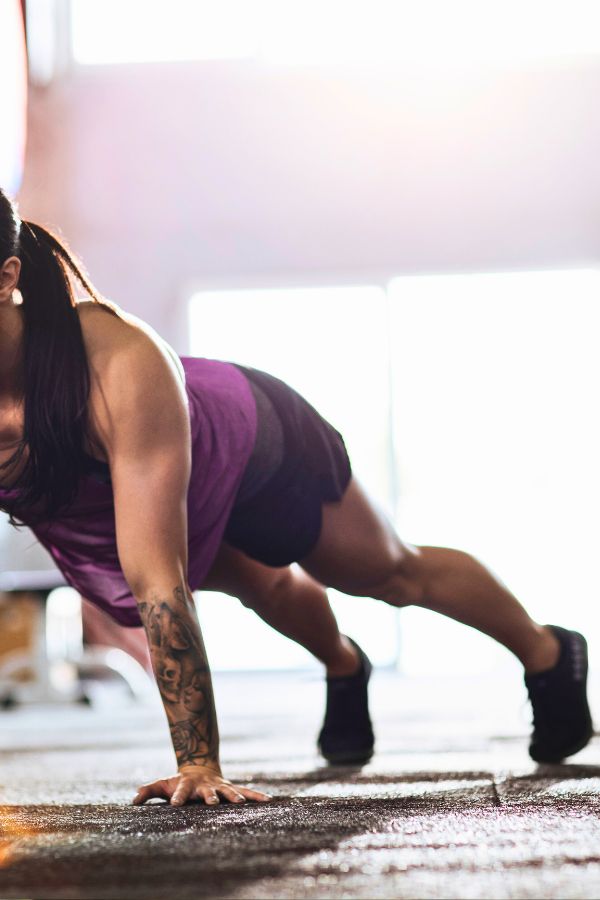 A person in a purple tank top and black shorts performs a plank indoors, sunlight streaming through large windows, as part of their Push Ups Workout routine.