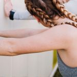 Two people with braided hair and athletic clothing lean against a white wall, performing Wall Push Ups as they stretch or warm up with their arms extended.